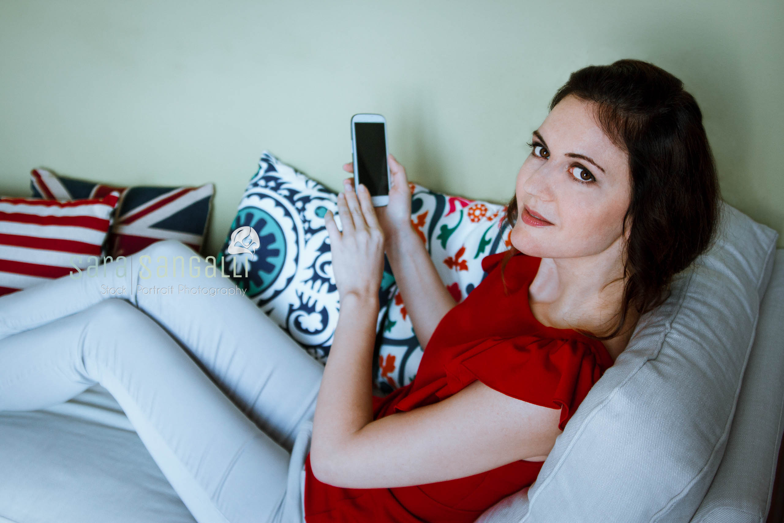Brunette holding smartphone while sitting on a couch and looking at camera