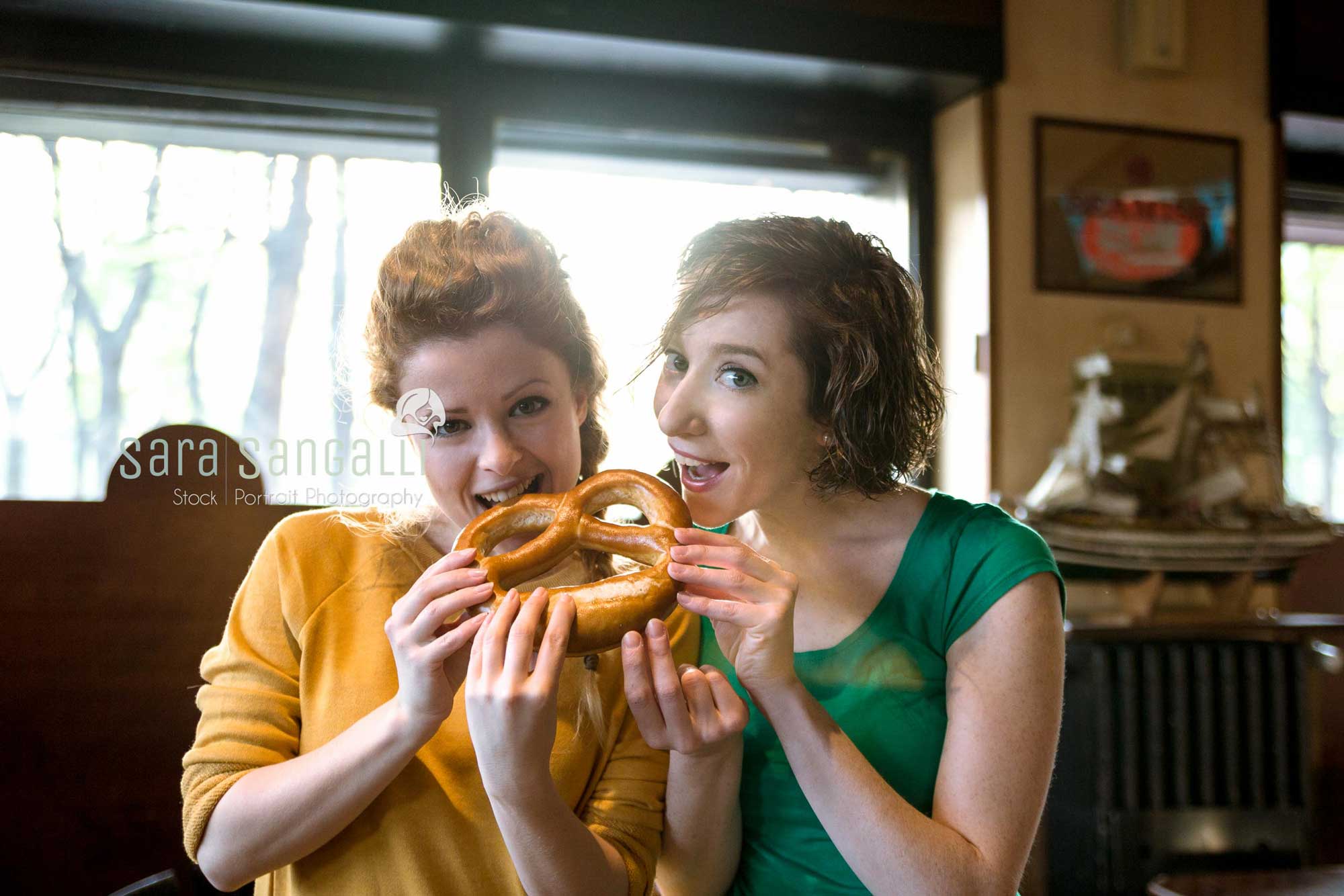 Two girlfriends eating pretzel, looking at camera indoor setting